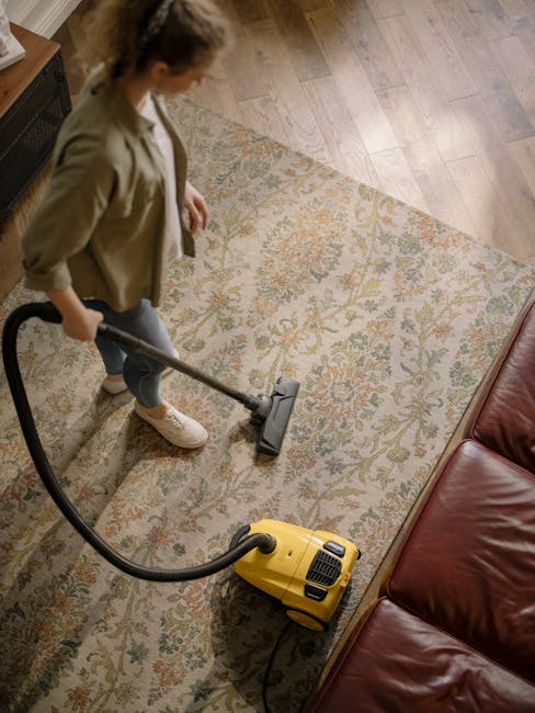 A person holds a yellow portable industrial air mover, positioned on a patterned area rug with floral motifs, to assist in surface drying or deep cleaning in a residential or commercial space. The roo