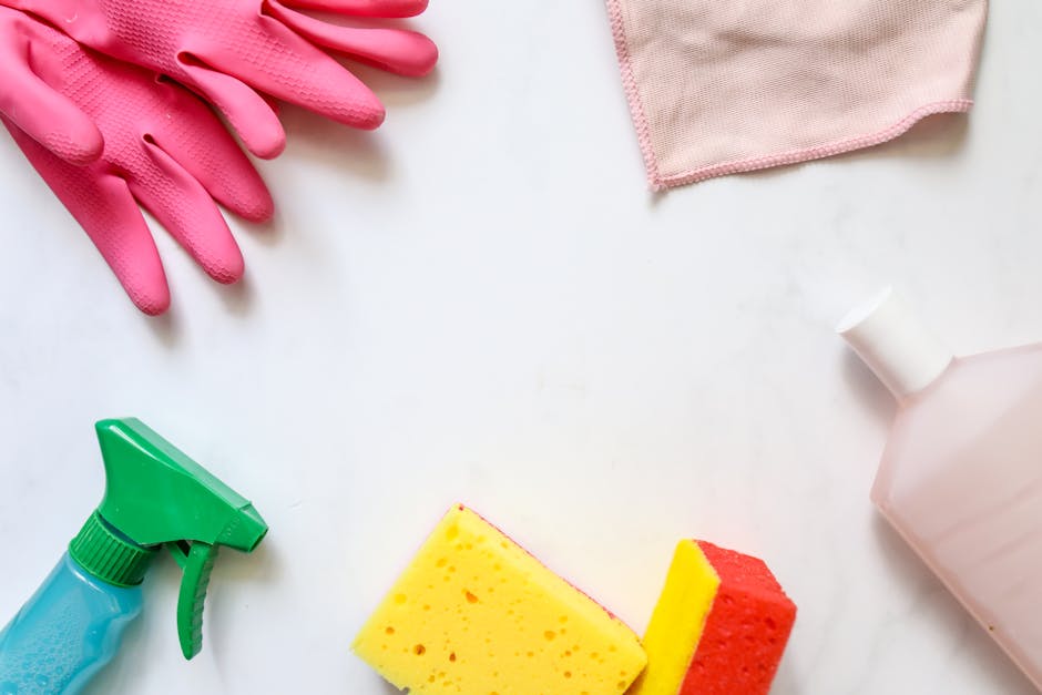 A flat lay of cleaning supplies arranged on a white surface, including a pair of pink rubber gloves, a pink cleaning cloth, a green spray bottle, a yellow and red sponge, and a white spray bottle, illustrating tools used for surface cleaning, deep cleaning, and sanitisation in domestic spaces. The arrangement reflects a focus on hygiene and thorough cleaning processes, consistent with residential and commercial cleaning standards emphasized by Kensington Cleaners for end of tenancy cleaning near Earls Court, W8 flats.