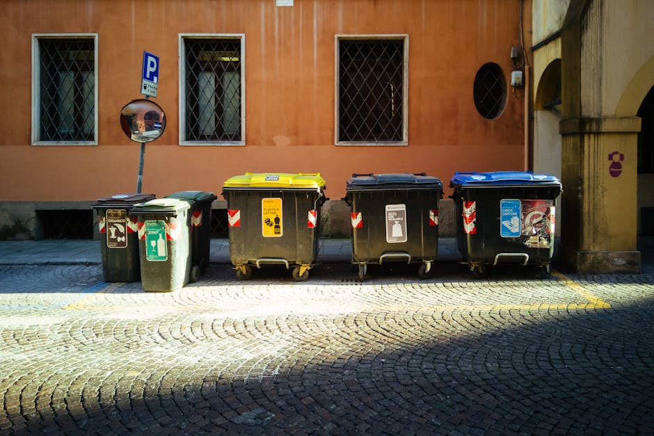 A row of sealed yellow and white garbage bags filled with household waste, placed on a cobblestone pavement against a white textured wall. The bags are made of plastic, some showing labels and black t
