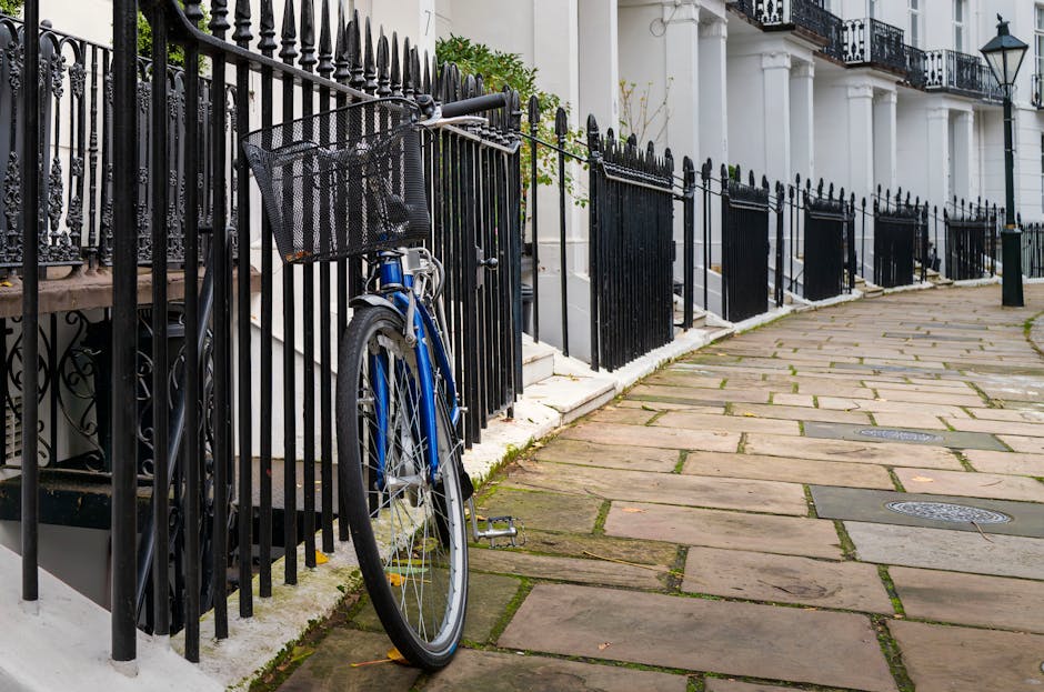 A blue bicycle with a black basket attached to the front handlebars is parked against a black metal fence along a paved sidewalk. The fence features pointed finials and runs parallel to a row of white residential buildings with black iron balconies and classic lamp posts. The sidewalk consists of large, uneven stone slabs, some with visible moss or dirt in the joints. The scene is outdoors during daytime, with natural light illuminating the area, and the overall appearance suggests a well-maintained urban residential setting. This image highlights surface cleaning and maintenance in a high street setting, consistent with the services offered by Kensington Cleaners, as referenced on the Kensington High Street guide to local domestic cleaning services page.