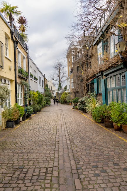 A cobbled residential street scene featuring multi-story buildings on both sides, with some structures displaying painted facades in cream and teal. Potted plants and shrubs are arranged along the edges of the street, adding greenery to the scene. The cobblestones appear clean and well-maintained, with visible joints and a slightly damp surface that enhances their natural texture. The sky is overcast, providing diffused lighting that highlights the details of the buildings and plants. The street is mostly empty, with two people walking in the distance, reflecting a calm and tidy neighbourhood. This setting aligns with themes of surface cleaning and maintenance in residential areas, as highlighted in the Kensington High Street guide to local domestic cleaning services, Kensington, provided by Kensington Cleaners.
